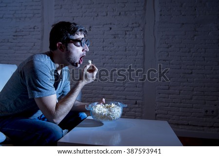 young television addict man sitting on home sofa watching TV and eating popcorn wearing funny nerd and geek glasses looking mesmerized enjoying movie sitcom or live sport at night