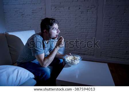 young television addict man sitting on home sofa watching TV and eating popcorn looking mesmerized enjoying movie sitcom or live sport at night