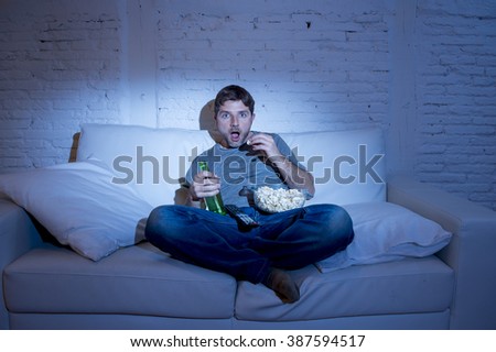 young television addict man sitting on home sofa watching TV eating popcorn and drinking beer bottle looking mesmerized enjoying movie sitcom or live sport at night