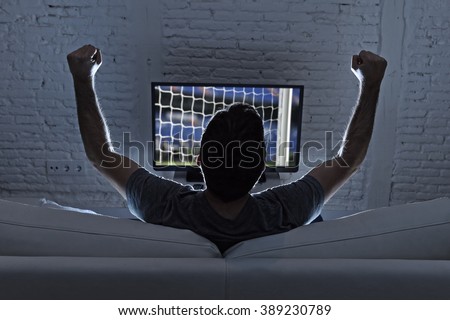 back view of young man home alone watching soccer or football game in television enjoying and celebrating goal and victory gesturing with fist sitting on the sofa happy and excited
