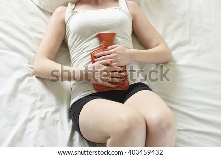young beautiful woman suffering stomach cramps on belly holding with hands hot water bottle against tummy in period pain lying on bed at home in menstruation hurt concept