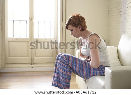 young beautiful red hair woman holding hurting belly suffering stomach cramp and period pain sitting on home couch in painful face expression female menstruation concept