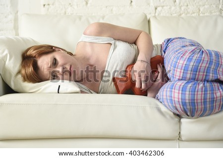 young beautiful red hair woman holding hot water bottle in a hurting tummy suffering stomach cramp and period pain lying on home couch in painful face expression female menstruation concept