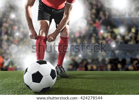 close up legs of football player with ball wearing black shoes adjusting his red sock standing on stadium pitch with background flashes and light flares