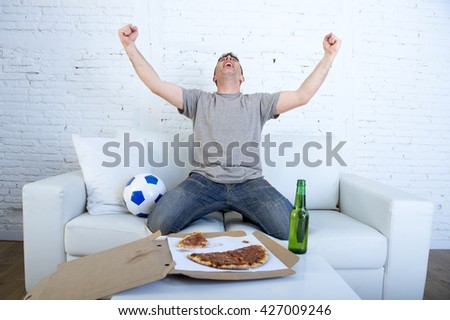 young man watching football game on television celebrating goal crazy happy jumping on sofa couch at home with ball beer bottle and pizza looking excited and cheerful