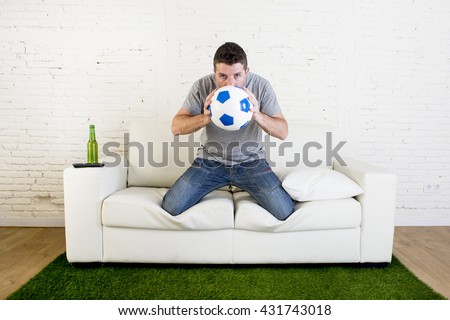 crazy football fan cheering watching television soccer match suffering stress nervous and excited kissing the ball hoping luck on sofa couch with grass carpet emulating stadium pitch