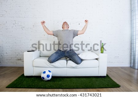 crazy football fan cheering happy watching television soccer match celebrating scoring goal excited and euphoric in sofa couch with ball and  grass carpet emulating stadium pitch