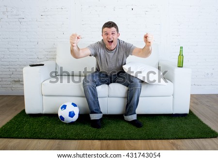 crazy football fan cheering happy watching television soccer match celebrating scoring goal excited and euphoric sitting on sofa couch with ball and grass carpet emulating stadium pitch