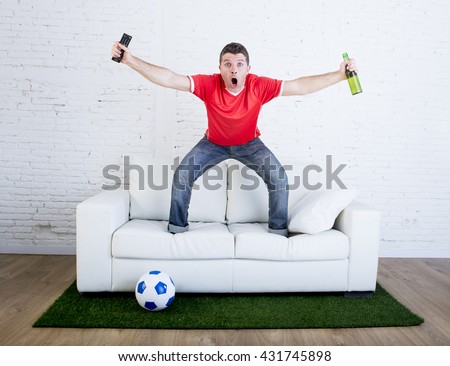 crazy football fan in red team jersey cheering happy watching television soccer match celebrating scoring goal excited and euphoric in sofa couch with ball on grass carpet emulating stadium pitch