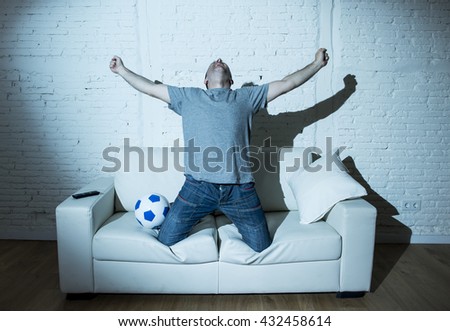 young man fanatic and crazy football fan watching television soccer match alone screaming happy celebrating scoring goal in glad in ecstasy with ball on home couch