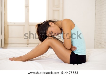 young beautiful hispanic woman in painful expression holding her belly suffering menstrual period pain lying sad on home bed having tummy cramp in female health concept