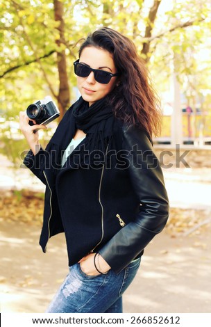 Outdoor fashion portrait of stylish photographer girl holding vintage retro camera, wearing trendy sunglasses and leather jacket. Lifestyle portrait bright toned colors.