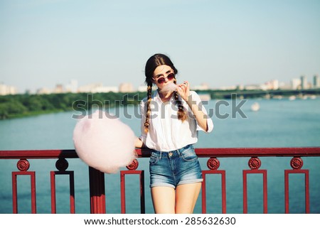 Lifestyle. Young happy hipster woman eating sweetened cotton candy, amazing view of the  river and the city from the bridge.