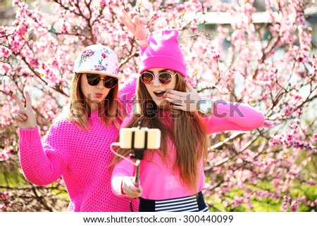 Closeup fashion lifestyle portrait of two young pretty beautiful friends in the lush spring garden on a sunny day. Cheerful friends, having fun together, joy and making selfie with self monopod.