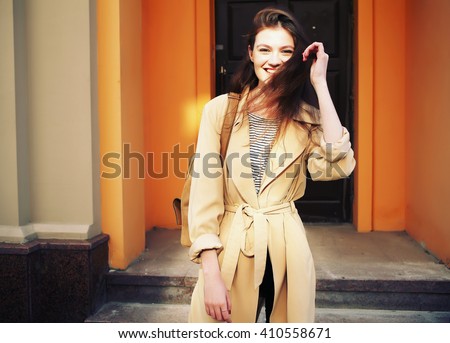 Young stylish hipster girl wearing modern trench coat walking in urban city smiling happy. Caucasian female model in her 20s.