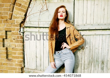 Street fashion concept - closeup portrait of a pretty hipster girl. Wearing brown leather jacket and high waisted jeans. Beautiful autumn woman with red libs and curly hair. Artsy bohemian style.