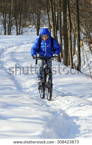 Extreme winter cyclist riding mountain bicycle with effort on rural track among snowbanks near snowy beautiful forest in sunny cold day