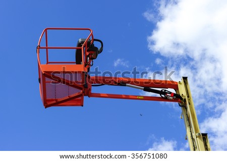 Hydraulic lift platform with bucket of construction vehicle painted in orange and beige colors with white clouds and blue sky on background