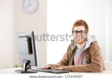 Young self-confident office worker with nerdy glasses smiling at you while working on his computer