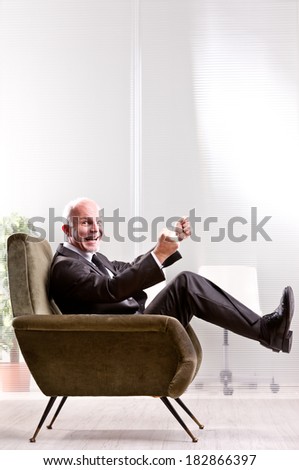 Cheering and exulting mature businessman on an armchair in an office laughing and looking at you