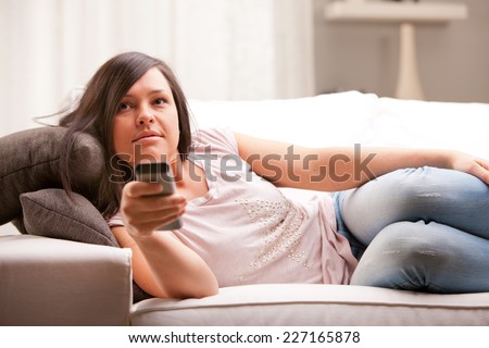 young woman watching tv on her sofa in her living room changing programme using a gray remote control