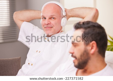 man smiling while listening to music in a living room while his friend does something else