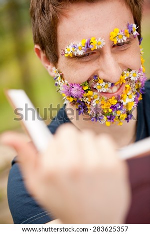 full nature immersion for a smiling hipster young man reading a book having his face covered with flowers instead of his beard and eyebrows (vertical)