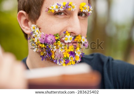 young man with his face flower-covered on his beard and eyebrows reading and staring at you very self-confident