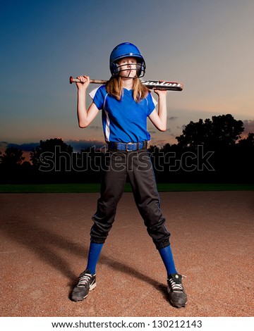 Girl in blue softball uniform at night with bat across shoulders.