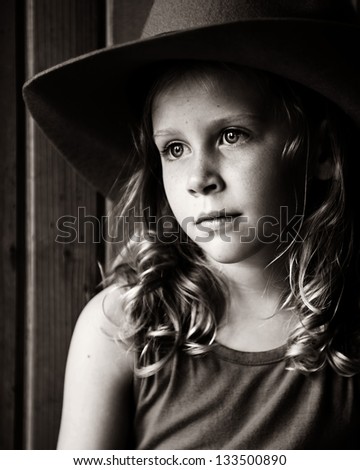 Little girl sitting in barn with light on her face, wearing cowboy hat and looking wistful