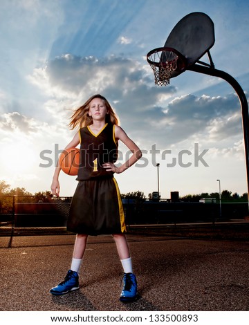 Little girl in black and gold basketball uniform standing next to playground hoop with ball, dramatic sky