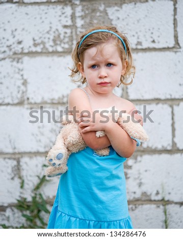 Scared little girl with blue eyes wearing blue dress and clutching stuffed bunny against white brick wall