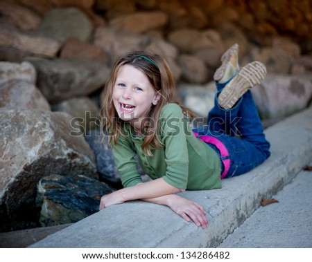 Laughing little girl with missing teeth, in green shirt and jeans lying on concrete curb with rocks behind her