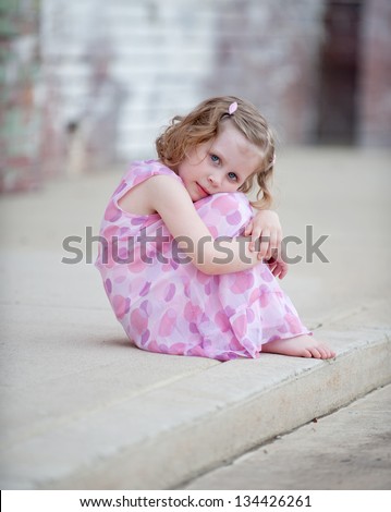 Cute little barefoot girl in pink dress sitting on curb hugging her knees