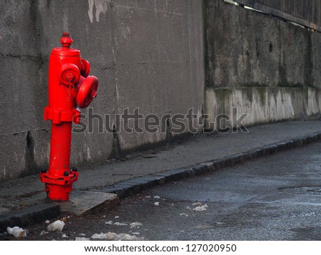 A red hydrant on a street. Both the road and the background wall are very gray contrasting with the fire hydrant. Some dirt on the hydrant and also some show around
