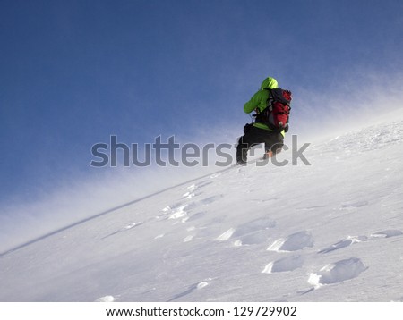 A climber making a path towards the mountain summit in deep snow blown by high winds
