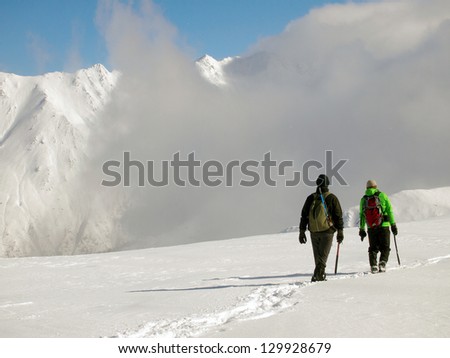 Two climbers with ice axe and crampons moving on snow, with summits and clouds in the background