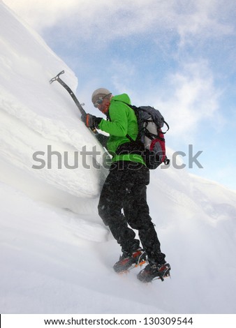 A climber with ice axes and crampons climbing a snow cornice in very high wind conditions. The wind is blowing snow and ice particles all over the climber almost covering him
