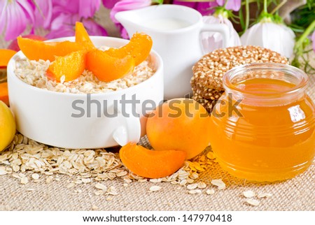Oatmeal with apricot in the bowl, honey, oatmeal cookies with sesame, milk jug and flowers