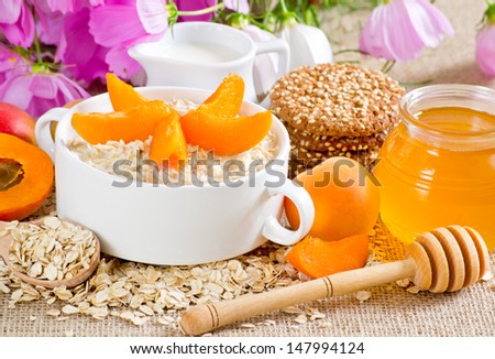 Oatmeal with apricot in the bowl, honey, oatmeal cookies with sesame, milk jug and flowers