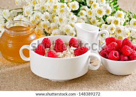 Oatmeal with raspberries in the bowl, honey, milk jug and flowers on the sackcloth