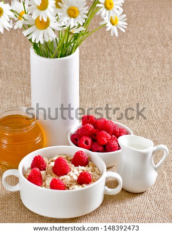 Oatmeal with raspberries in the bowl, honey, milk jug and flowers on the sackcloth