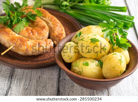 Young boiled potatoes with dill in the bowl, sausage on the plate, parsley and onions on the wooden board