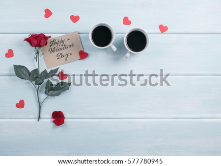 Red rose and two coffee cups with hearts and paper greeting or invitational card for Valentine day with place for text on background of shabby wooden planks. Copy space.