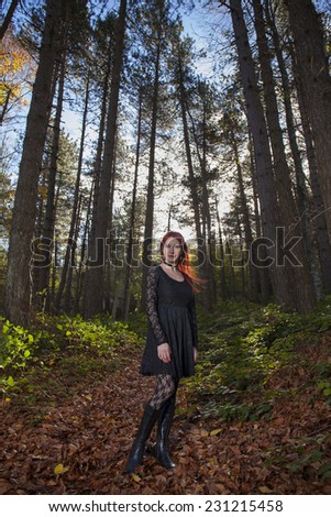 Beautiful young girl with red hair in gothic clothing in a forest with tall trees  and blue sky background