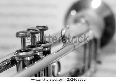 Trumpet as seen from behind with blurred out sheet music as background and first keys in focus, black and white.