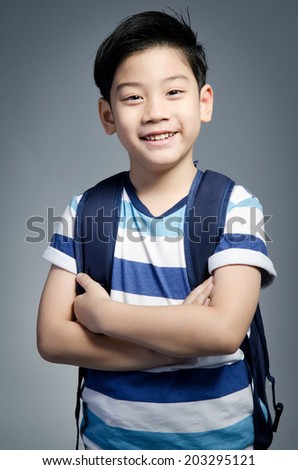 Little Asian child standing with a kit bag slung over his shoulder on gray background .