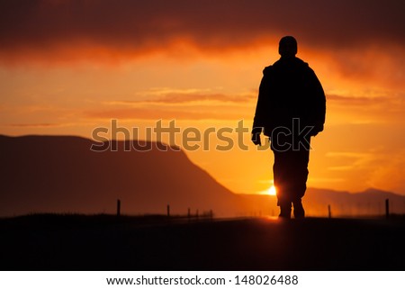 A Man Walking Along The Road, Backlit At Sunset, Among The Mountains, The Clouds