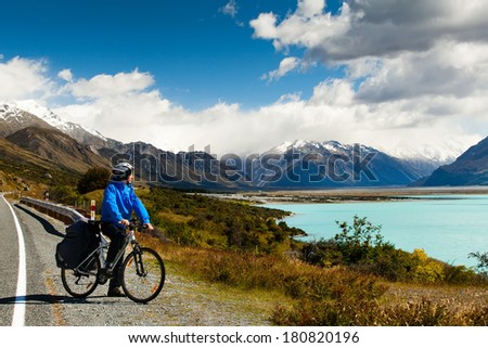 cyclist stands on the winding asphalt mountain road near Lake Pukaki view from Glentanner Park Centre near Mount Cook, on a background of blue sky with clouds, snowy Southern Alps.
