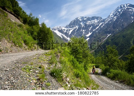 A man rides a bicycle on a mountain gravel road. Beautiful view of alpine snowy meadows in the village Zhabeshi. Caucasus mountains. Upper Svaneti, Georgia.